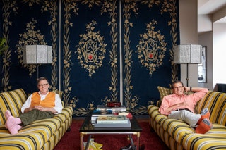 Three blue decorative panels on far wall behind two matching striped yellow sofas on which two men are seated wearing...