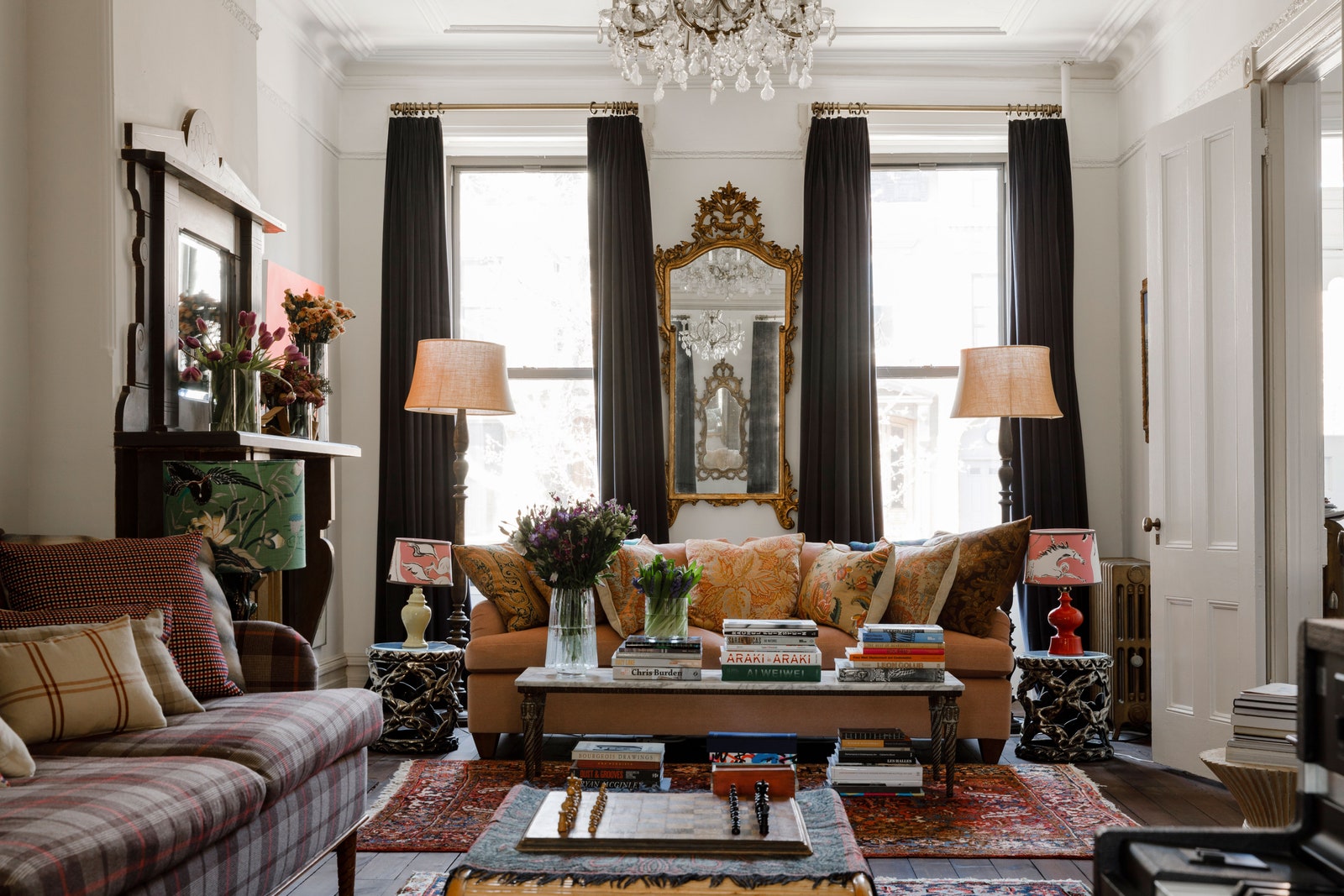 Living room of a New York brownstone home with a gilded mirror flanked by two tall windows chandelier hanging from...