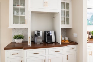 Coffee machines like an espresso machine or a Keurig are tucked away inside the cabinet in this space designed with...